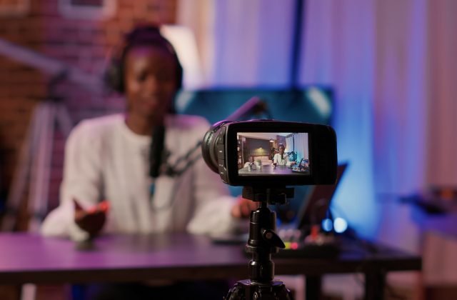 Selective focus on african american woman influencer talking in front of digital camera recording vlog sitting at desk in home studio. Online radio host broadcasting live talk show late at night.